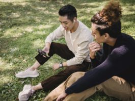 From above content young ethnic male friends wearing casual outfits surfing internet on modern mobile phone and sitting on grass in sunny park